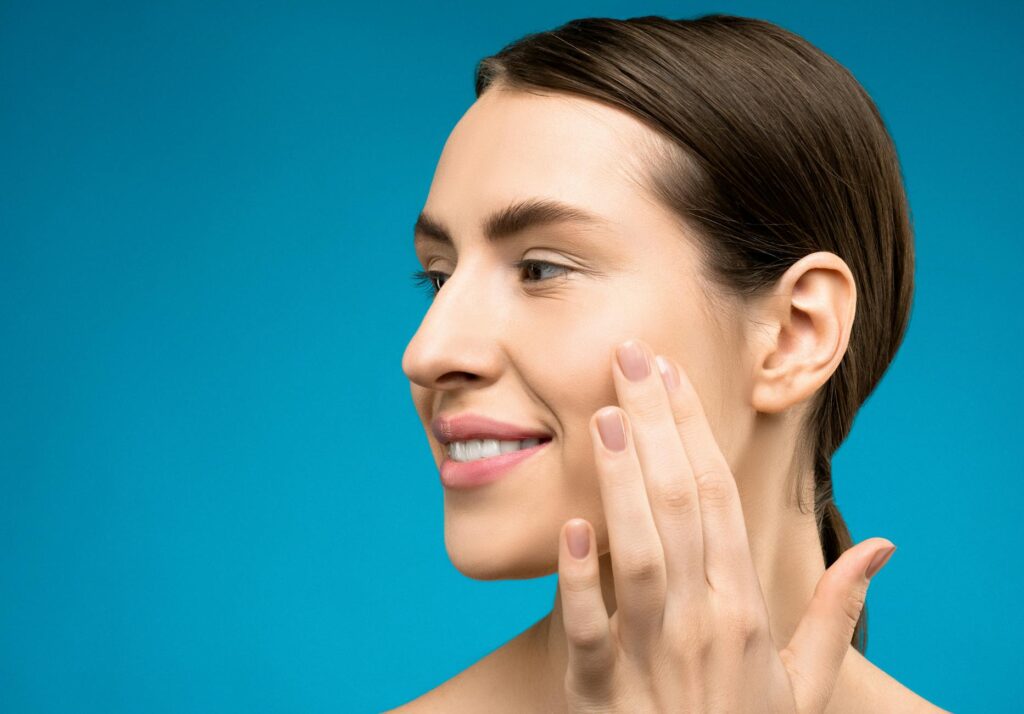 A close-up portrait of a smiling adult Caucasian woman touching her face against a blue background.
