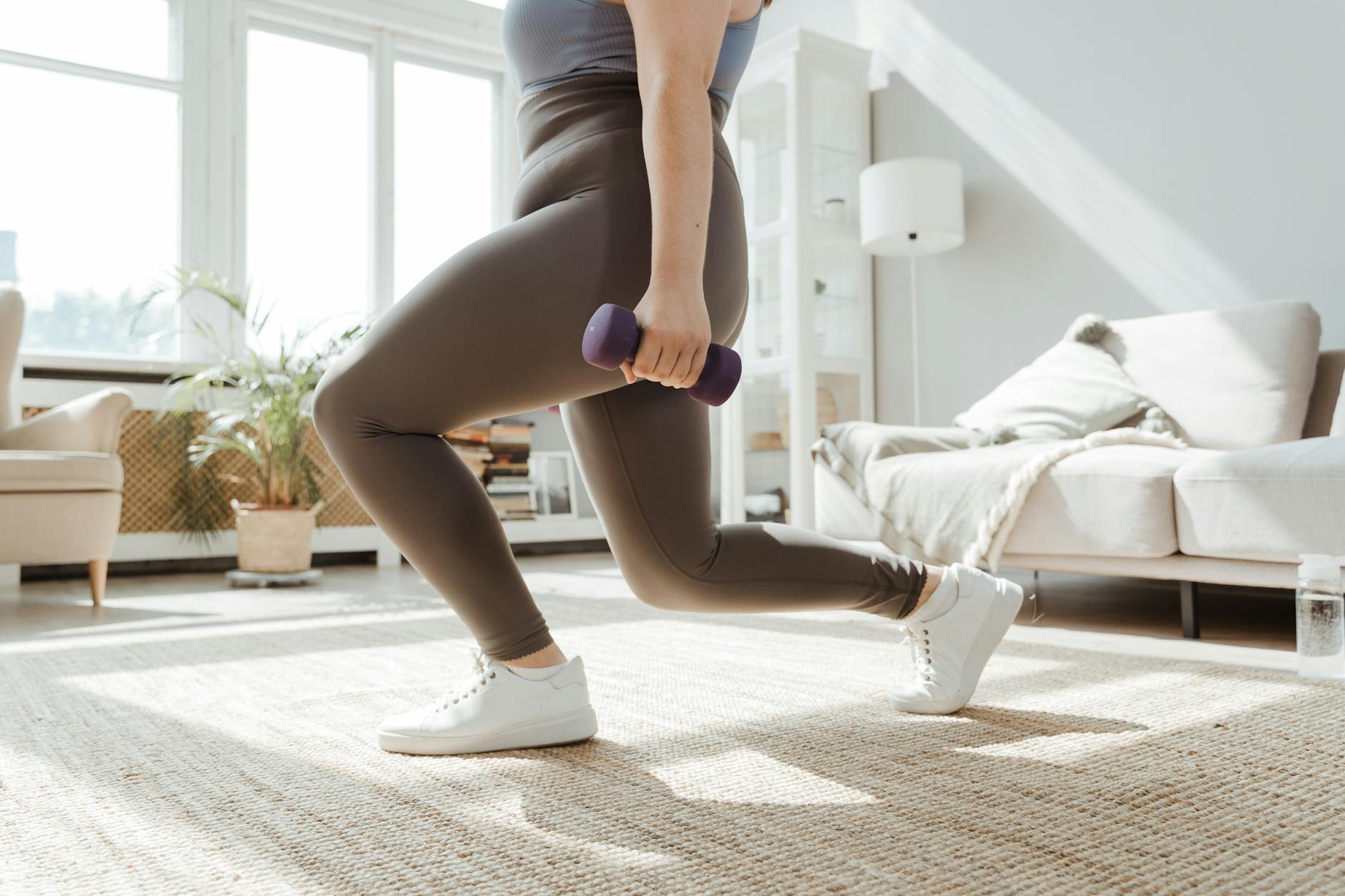 A woman in active wear performs a lunges exercise with dumbbells indoors.