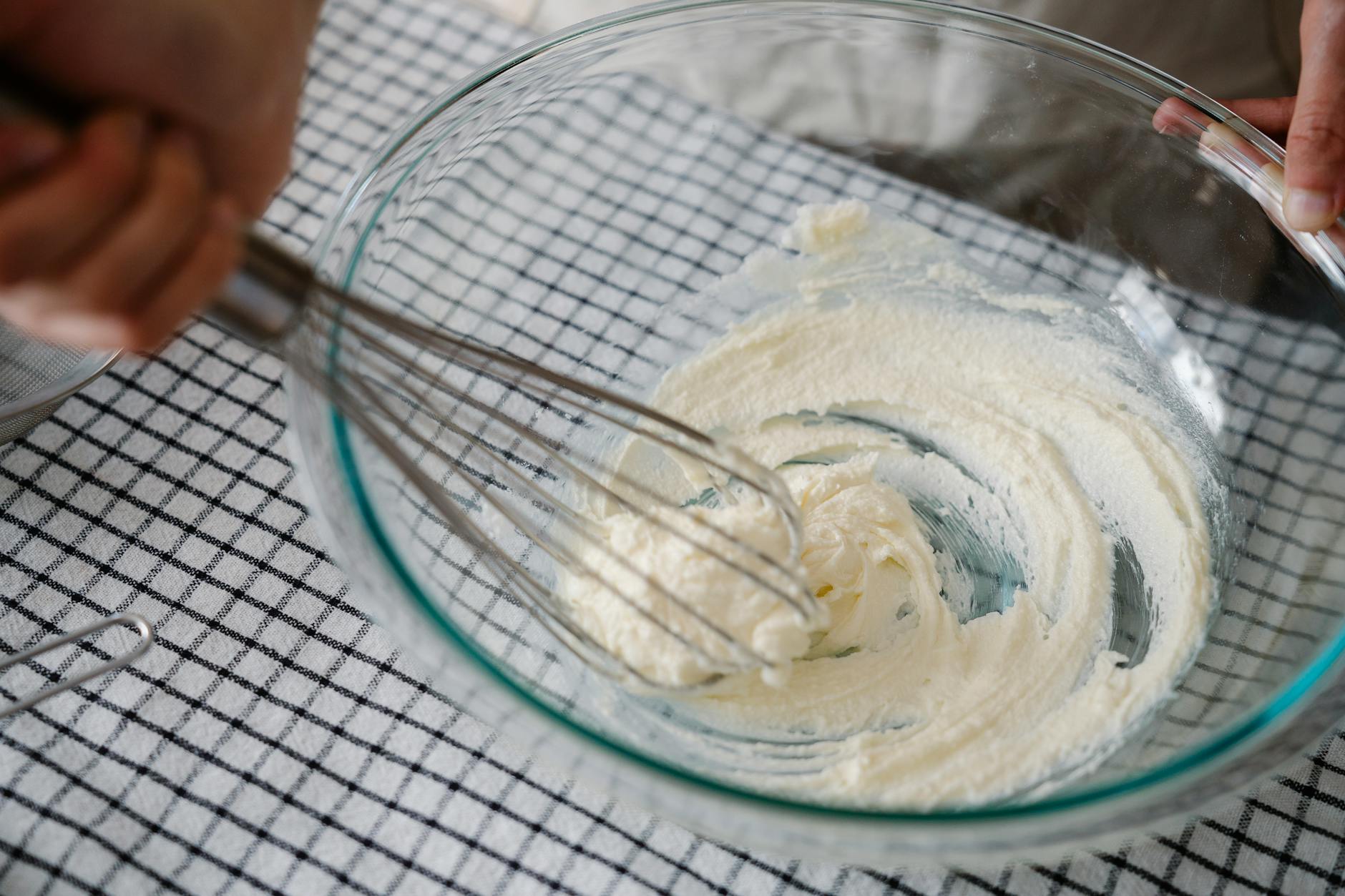 A person whisking cream in a glass bowl on a checkered tablecloth, capturing the art of baking.
